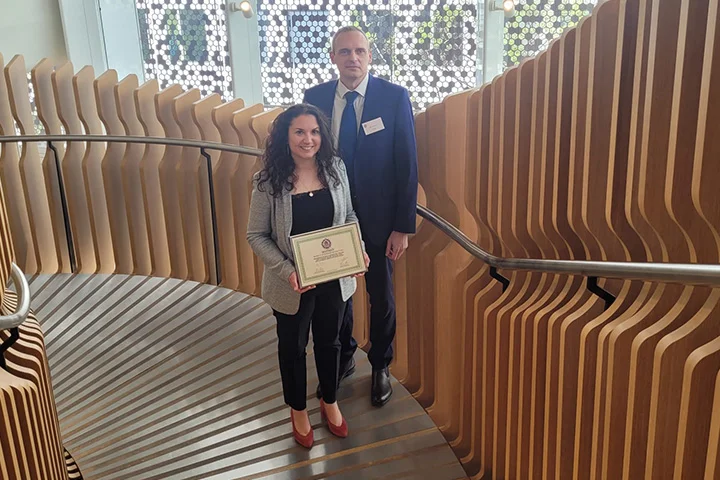 Deux personnes souriantes posent sur un escalier design en bois et métal. La femme tient un certificat.