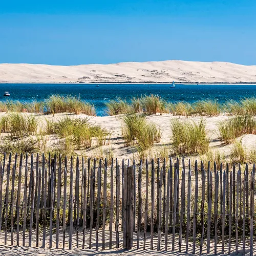 Paysage littoral ensoleillé montrant des dunes de sable et une clôture en bois face à une étendue d'eau bleue.